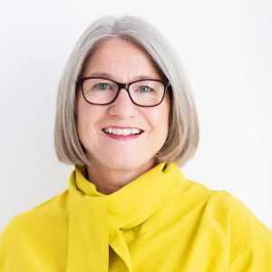 head shot of Marianne Heartly bobbed blonde hair wearing glasses ad a bright yellow jacket